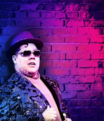 Person wearing a purple hat, sunglasses, and a sequined jacket poses in front of a brick wall illuminated with purple and pink lighting.