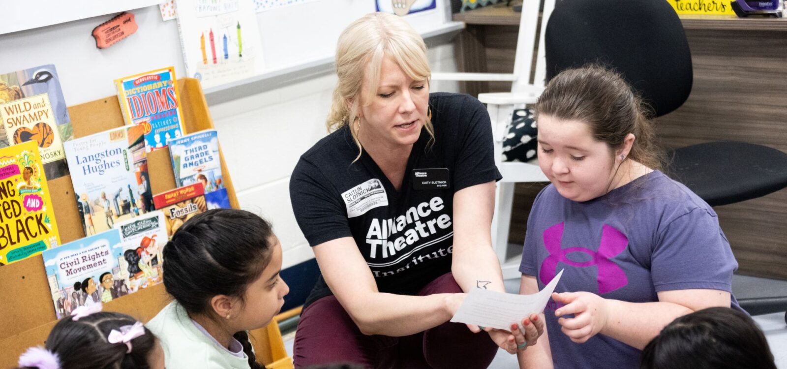 A woman in an "Alliance Theatre" shirt helps a young girl with a paper while other children sit nearby in a classroom with books on display.