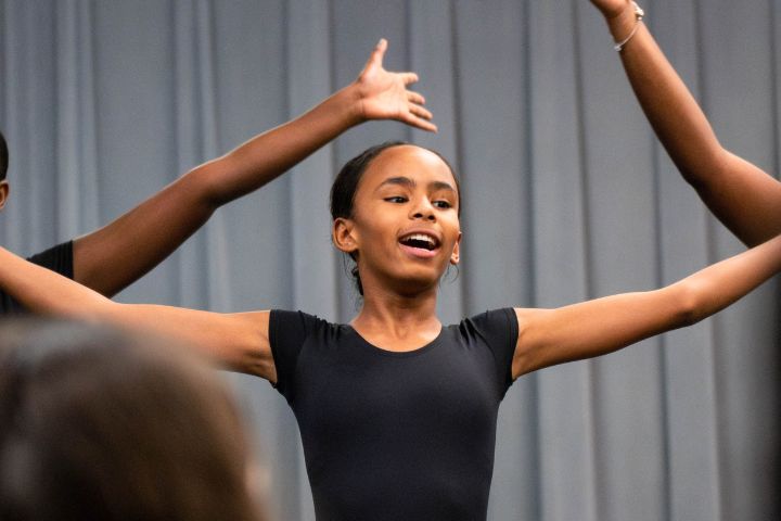 A young person in a black leotard performs a dance pose with arms raised, in front of a gray curtain background.