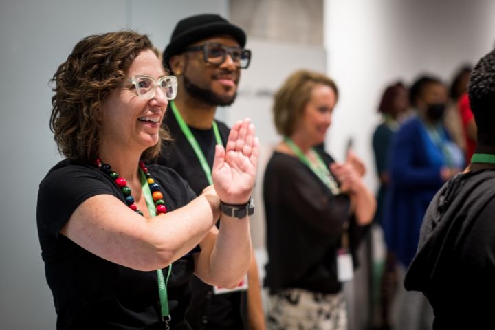 A group of people standing indoors, smiling and clapping, wearing name badges and green lanyards.