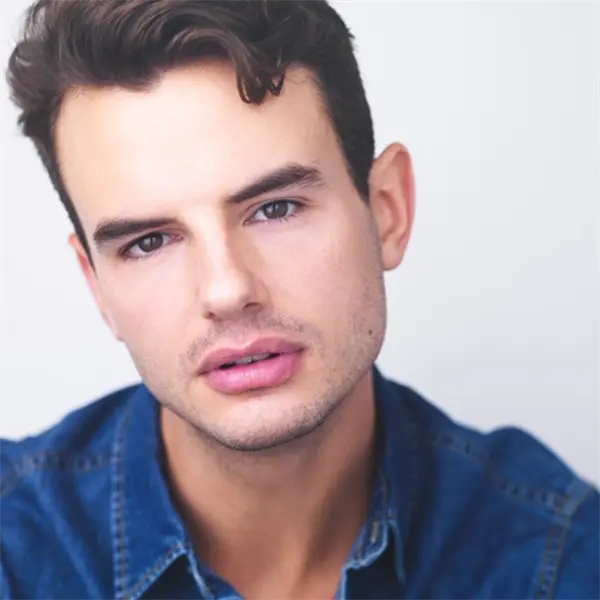 A man with short brown hair and light stubble wears a blue denim shirt and looks at the camera against a plain light background.