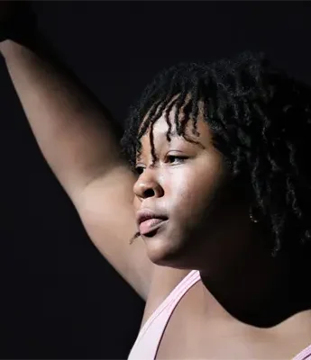 A person with short curly hair raises one arm against a dark background, illuminated by a focused light.