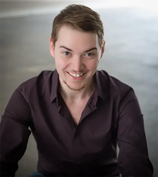 A young man with short light brown hair and a trimmed beard smiles at the camera while wearing a dark button-up shirt, seated in a well-lit indoor space.