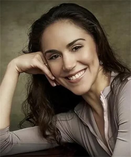 A woman with long, dark hair smiles at the camera, resting her head on her hand, wearing a light-colored long-sleeve top against a neutral background.