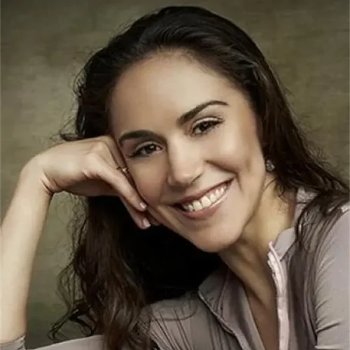A woman with long, dark hair smiles at the camera, resting her head on her hand, wearing a light-colored long-sleeve top against a neutral background.