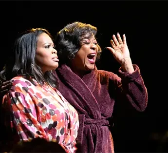 Two women wearing patterned and solid robes stand on stage under a spotlight, one looking ahead and the other gesturing with her hand while singing or speaking.