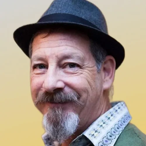 A middle-aged man with a goatee and mustache, wearing a gray fedora and a patterned collared shirt, smiles at the camera against a yellow background.