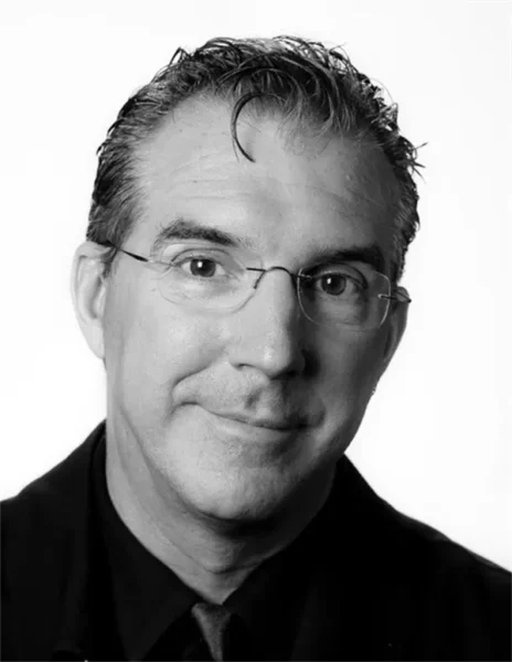 A man with short dark hair, wearing glasses, a dark jacket, and a tie, looks at the camera with a neutral expression against a plain background.