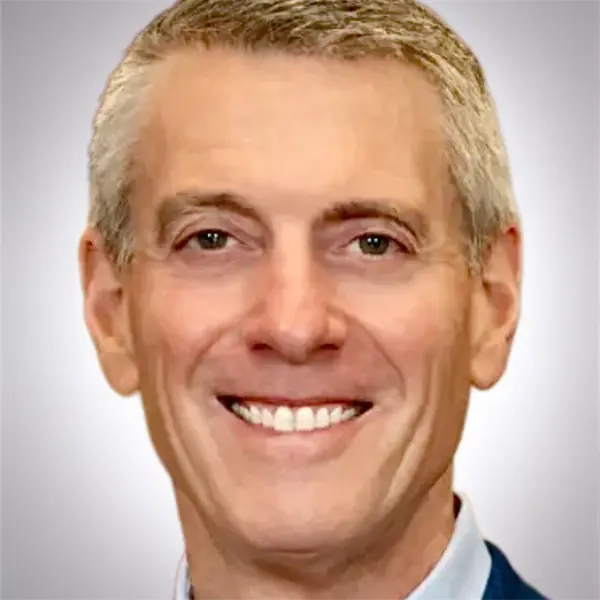A middle-aged man with short gray hair, light skin, and blue eyes is smiling at the camera against a neutral background.