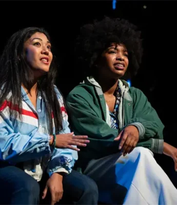 Two women sit side by side on stage under dramatic lighting, appearing engaged and expressive as they look into the distance.