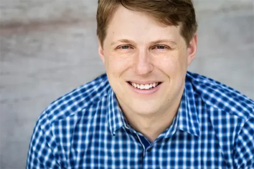 A man with light brown hair wearing a blue and white checkered shirt smiles at the camera in front of a neutral background.