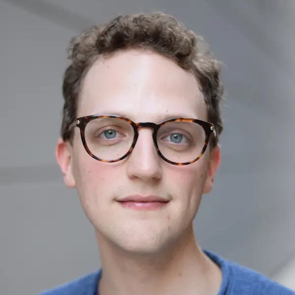 A person with short curly brown hair and glasses, wearing a blue shirt, stands in front of a light-colored background.