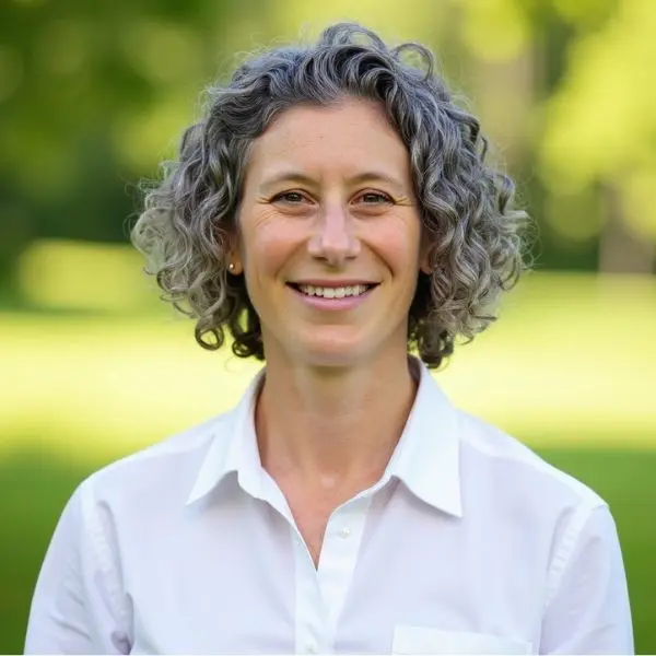 A person with short curly gray hair, wearing a white collared shirt, smiles outdoors with a blurred green background.