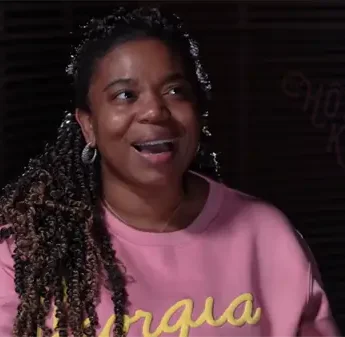 A woman with braided hair wearing hoop earrings and a pink sweatshirt smiles while sitting indoors against a dark background.