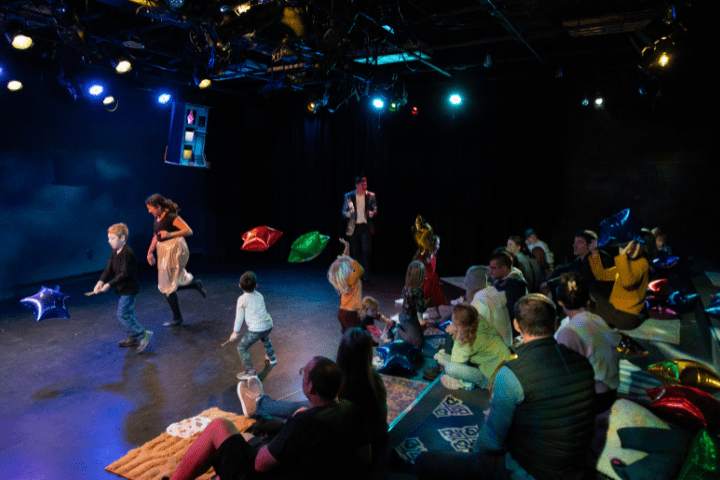 Children and adults watch and participate in a lively performance on a stage with colorful balloon decorations under stage lighting.