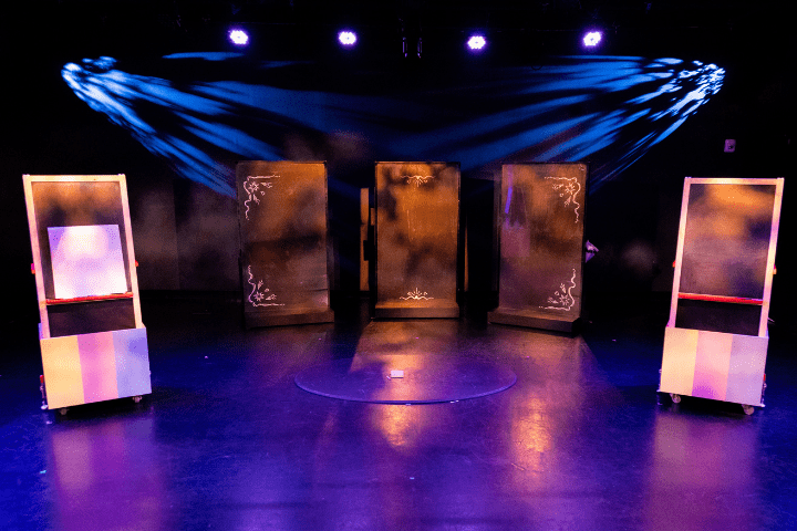 A stage set with five rectangular frames or panels under blue and purple lights, with a dark background and shadows cast on the floor and wall.
