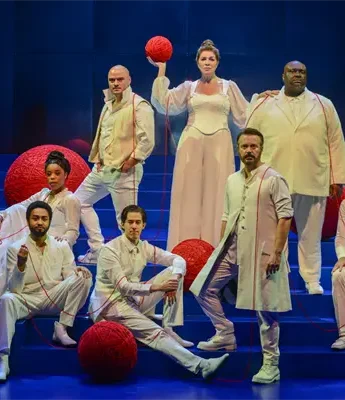 Eight actors in white costumes pose on a blue stage with large red yarn balls and threads, against a minimalist background.