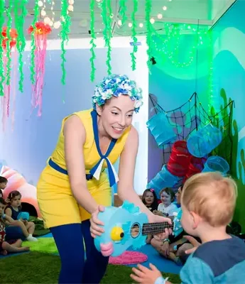 A performer in a yellow costume interacts with a young child in an underwater-themed room while other children and adults watch.