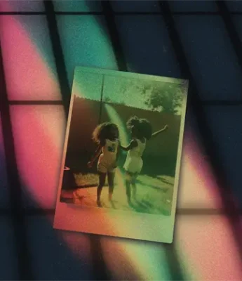 A Polaroid photo of two children outdoors is placed on a dark surface with colorful light streaks and grid shadows.