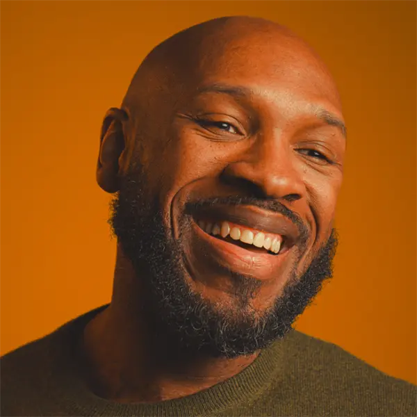 A man with a beard smiles warmly, facing the camera against an orange background, wearing a green sweater.