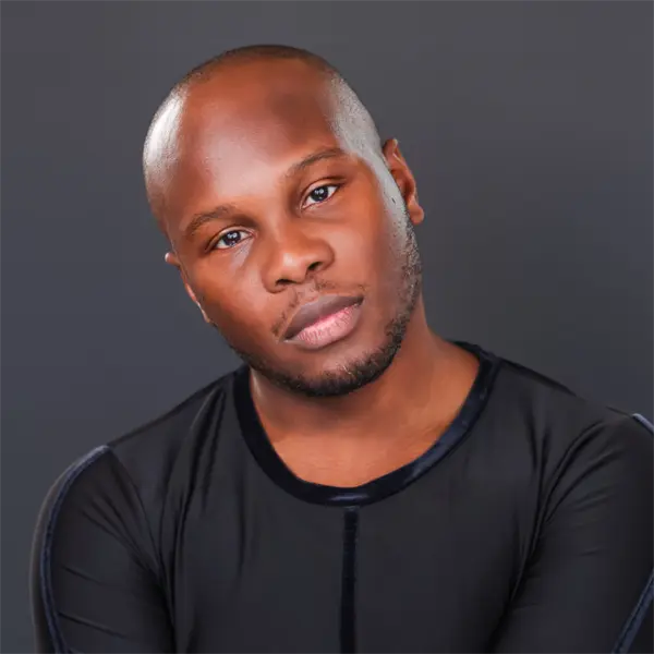 A person with a shaved head and short beard wearing a black shirt poses against a plain dark background.