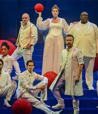 Eight actors in white costumes pose on blue steps, holding or surrounded by large red yarn balls, with red yarn connecting them.