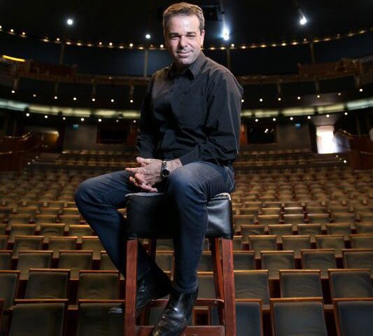 A man in a black shirt and jeans sits on a tall stool in the center of an empty theater, facing the camera. Rows of empty seats fill the background.