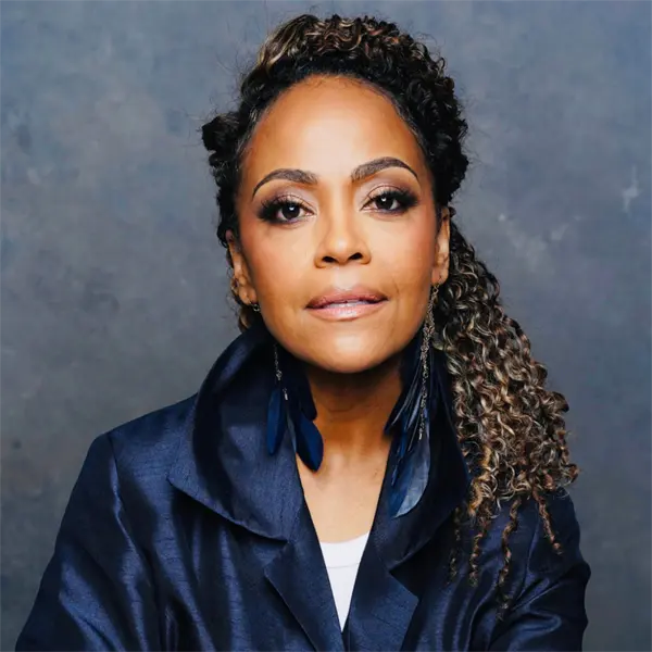 A woman with curly hair pulled to one side, wearing a dark blue jacket and statement earrings, poses against a gray background.