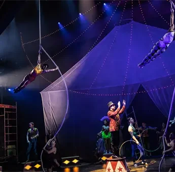 Performers in colorful costumes put on a circus-themed stage show, with acrobats suspended on ropes above a ringmaster and other entertainers under dramatic lighting.