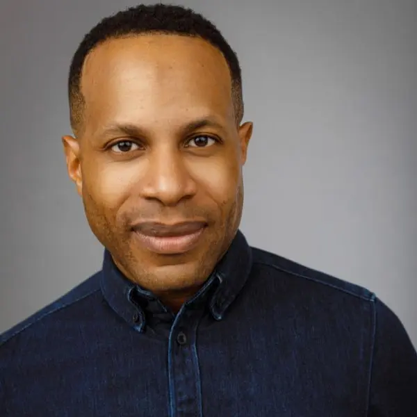 A man with short hair and a trimmed beard wearing a dark denim button-up shirt, standing against a plain gray background.