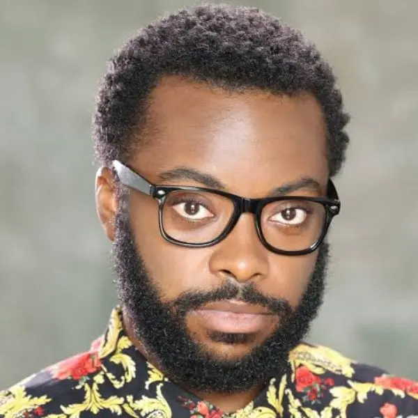 A man with short, curly hair, a full beard, and black-framed glasses, wearing a patterned shirt with yellow and red floral designs, looks directly at the camera.