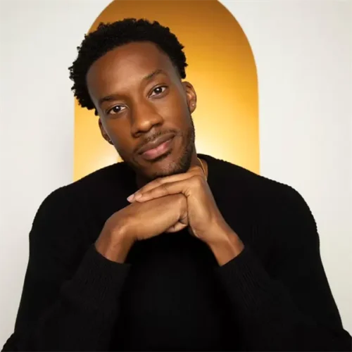 A man with short curly hair and a beard, wearing a black sweater, rests his chin on his hands in front of a golden arched background.