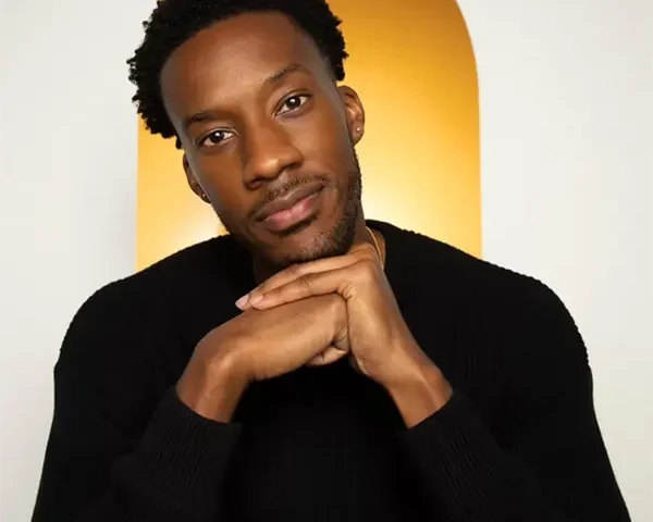 A man with short curly hair and a beard, wearing a black sweater, rests his chin on his hands in front of a golden arched background.