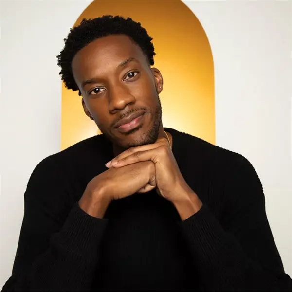 A man with short curly hair and a beard, wearing a black sweater, rests his chin on his hands in front of a golden arched background.