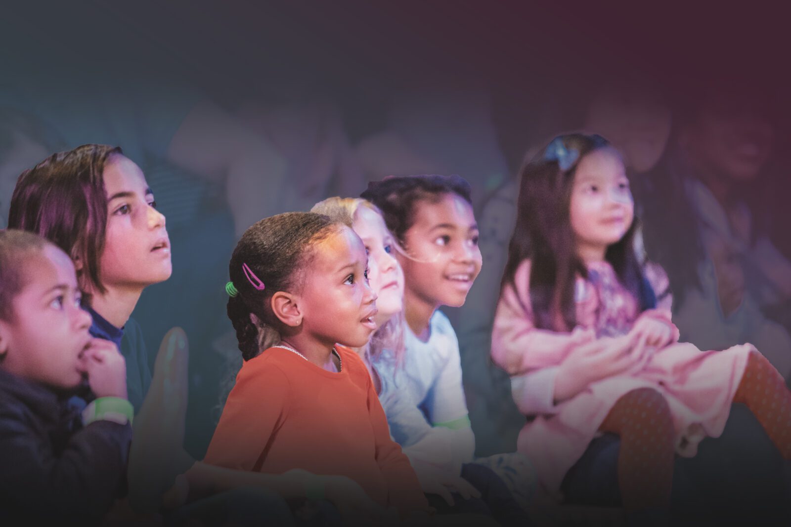 A group of young children sits closely together, looking ahead with interest and smiles, illuminated by soft, colorful lighting.
