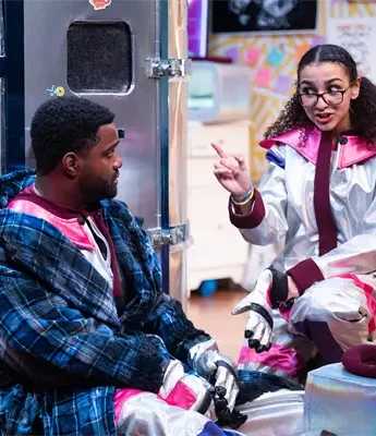 Two people in colorful costumes sit and talk next to a metallic fridge in a room decorated with bright patterns and sticky notes.