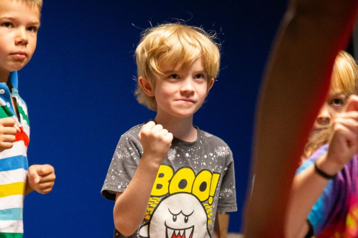 A young boy with blond hair stands with his fist raised, wearing a Boo! graphic t-shirt, among other children against a blue background.