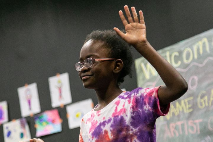 A child wearing glasses and a tie-dye shirt raises their hand, standing in front of a chalkboard and children's drawings displayed on a wall.