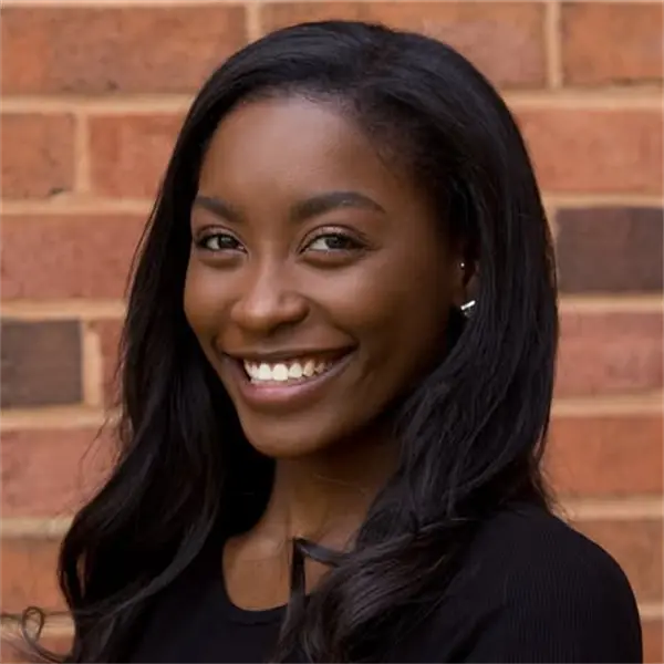 A woman with long dark hair smiles at the camera while standing in front of a brick wall, wearing a black top.