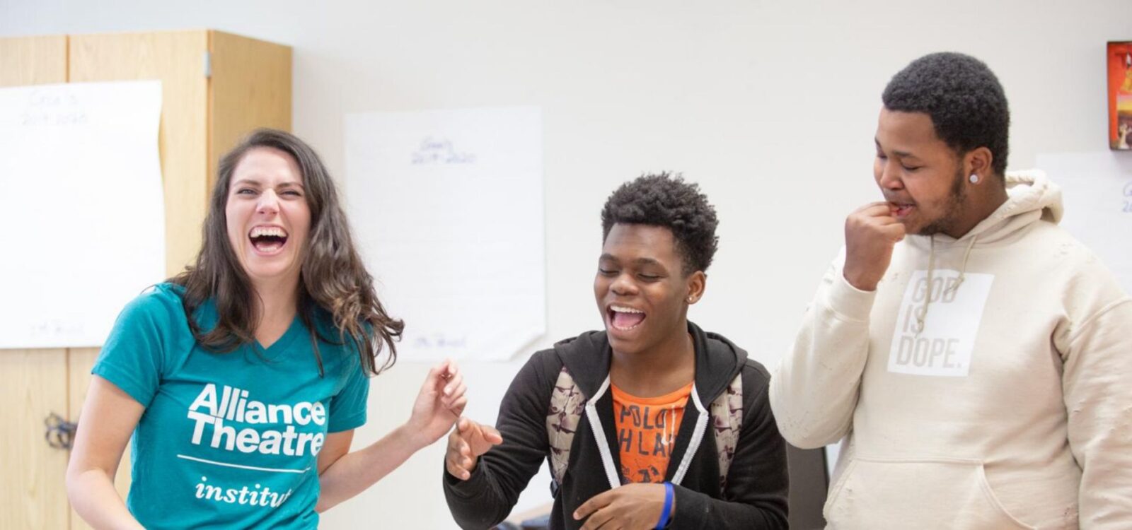 Three people stand indoors, smiling and laughing together. One wears an "Alliance Theatre" shirt, another an orange shirt, and the third a tan hoodie that says "God is Dope.