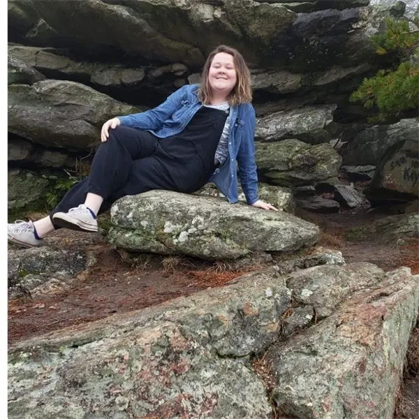 A person sits on a large rock formation outdoors, smiling at the camera, with rocky terrain and greenery in the background.