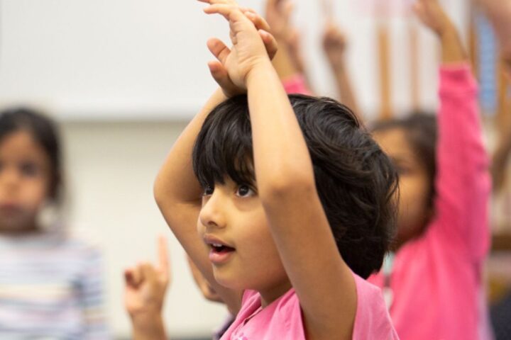 Several young children sit in a classroom with their hands raised, appearing engaged and attentive during a lesson or activity.