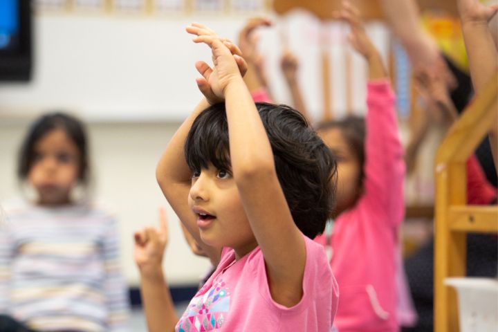 A young child in a classroom sits with hands raised above their head, looking attentive. Other children are seated around, also raising their hands.