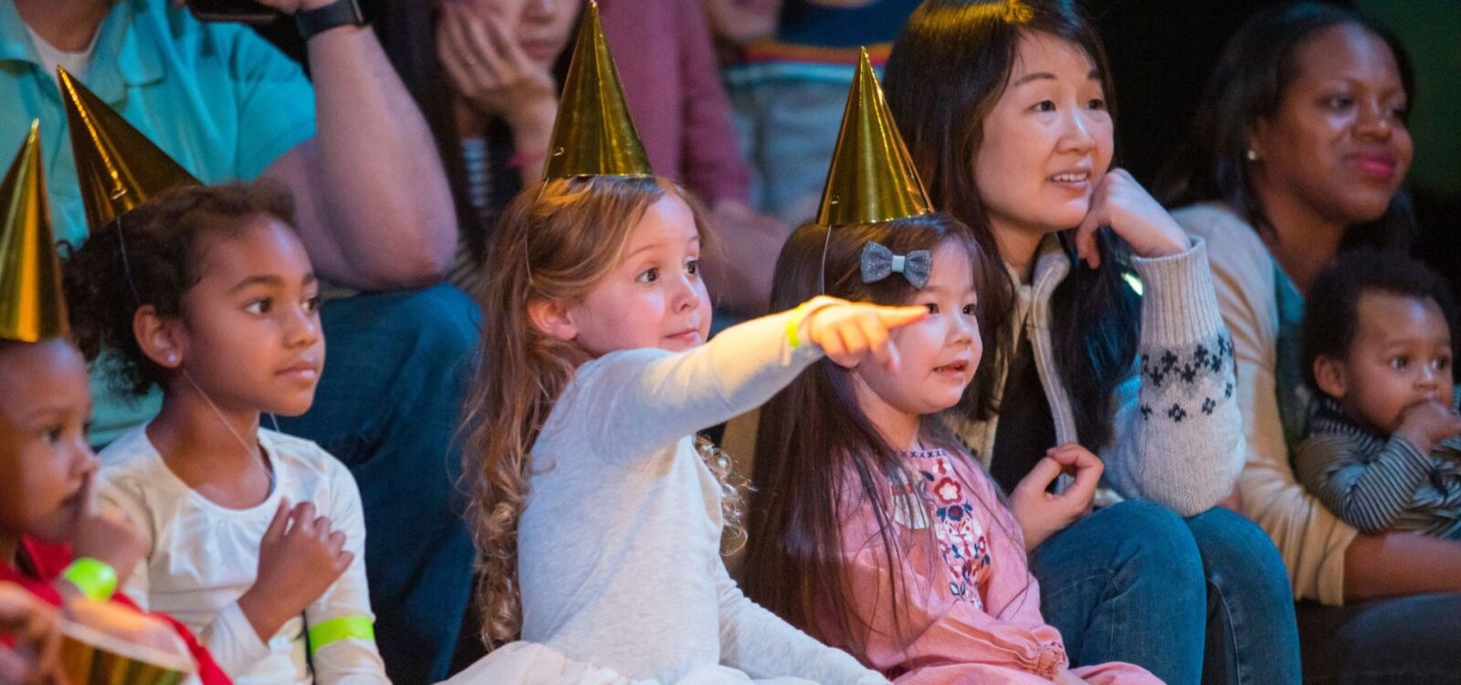 Children and adults wearing party hats sit closely together, watching something off-camera; one child is pointing excitedly.