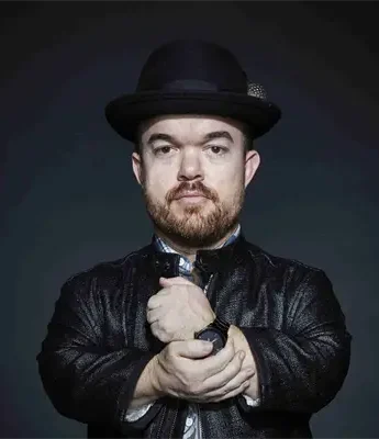 A man with a beard wearing a black bowler hat and black leather jacket stands against a dark background, holding one wrist with the other hand.