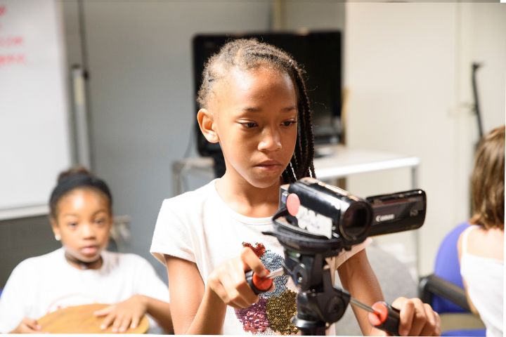 A girl adjusts a video camera on a tripod indoors, while another girl sits in the background.