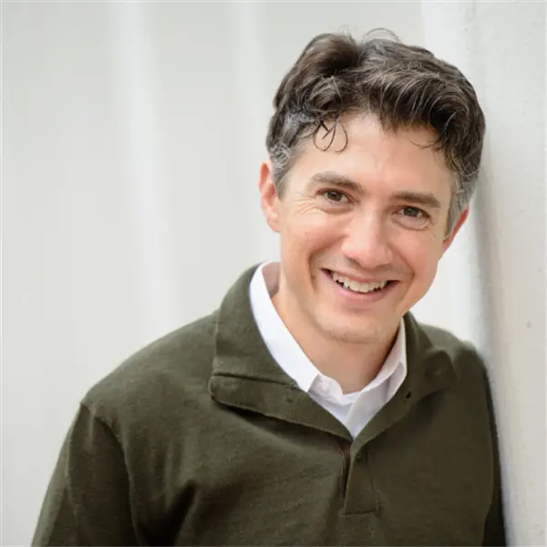 A man with short dark hair, wearing a green collared shirt over a white undershirt, smiles while leaning against a white wall.
