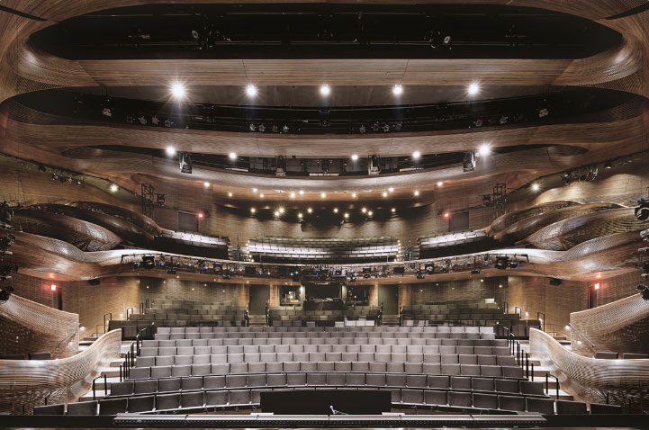 A modern, empty theater auditorium with multiple tiers of seating, wooden paneling, and illuminated stage lights.