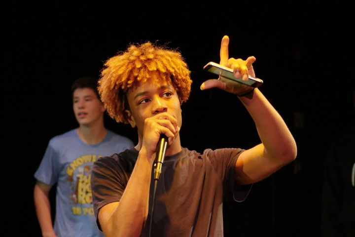A young man with light brown curly hair holds a microphone and a smartphone, gesturing with his hand, while another person stands in the background.