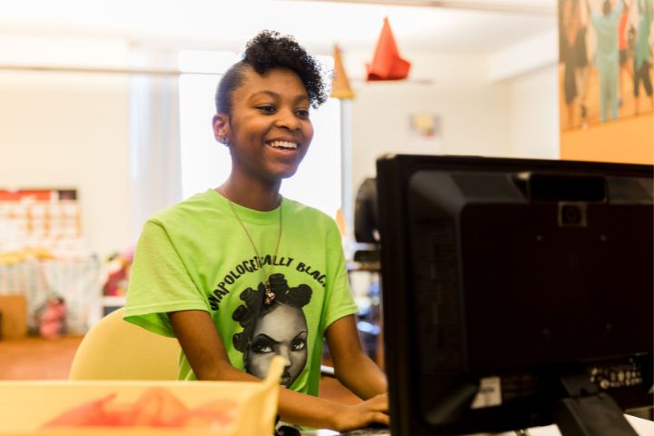 A person wearing a bright green t-shirt with a graphic design sits at a desk, smiling while using a desktop computer in a well-lit room.